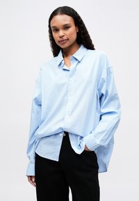 Camisa azul clara de botão com riscas verticais brancas, ajustada em tamanho oversized, mangas longas e colarinho clássico. Combinada com calças pretas.