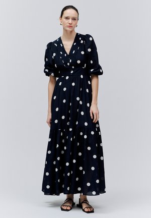 Kobieta stojąca w czarnej sukience maxi w białe groszki z bufiastymi rękawami, w zestawie z czarnymi płaskimi sandałami.