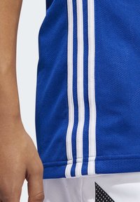 Närbild av en person som bär en blå sporttröja med tre vertikala vita ränder och vita shorts med svart prickigt mönster.