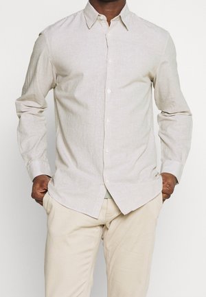 Homme portant une chemise beige rayée à manches longues et un pantalon beige, debout les mains dans les poches.