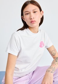 Hvit t-skjorte med kort erm og rosa logo på fronten, brukt sammen med lavendelbukser. Modellen har synlige tatoveringer på armen.