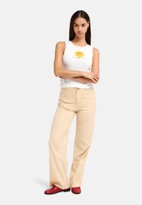 Débardeur blanc sans manches avec un motif jaune, pantalon beige ample à jambes larges, et chaussures rouges. La tenue présente un design décontracté et simple.