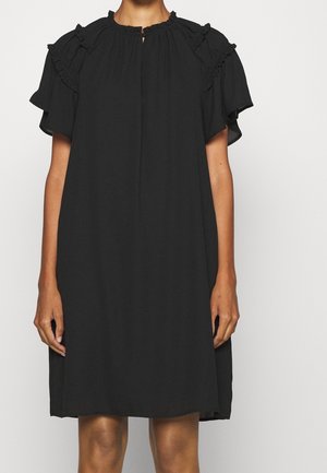 Kobieta w luźnej czarnej sukience z krótkimi falbaniastymi rękawami i dekoltem z wycięciem w kształcie klucza, stojąca na jednolitym tle.