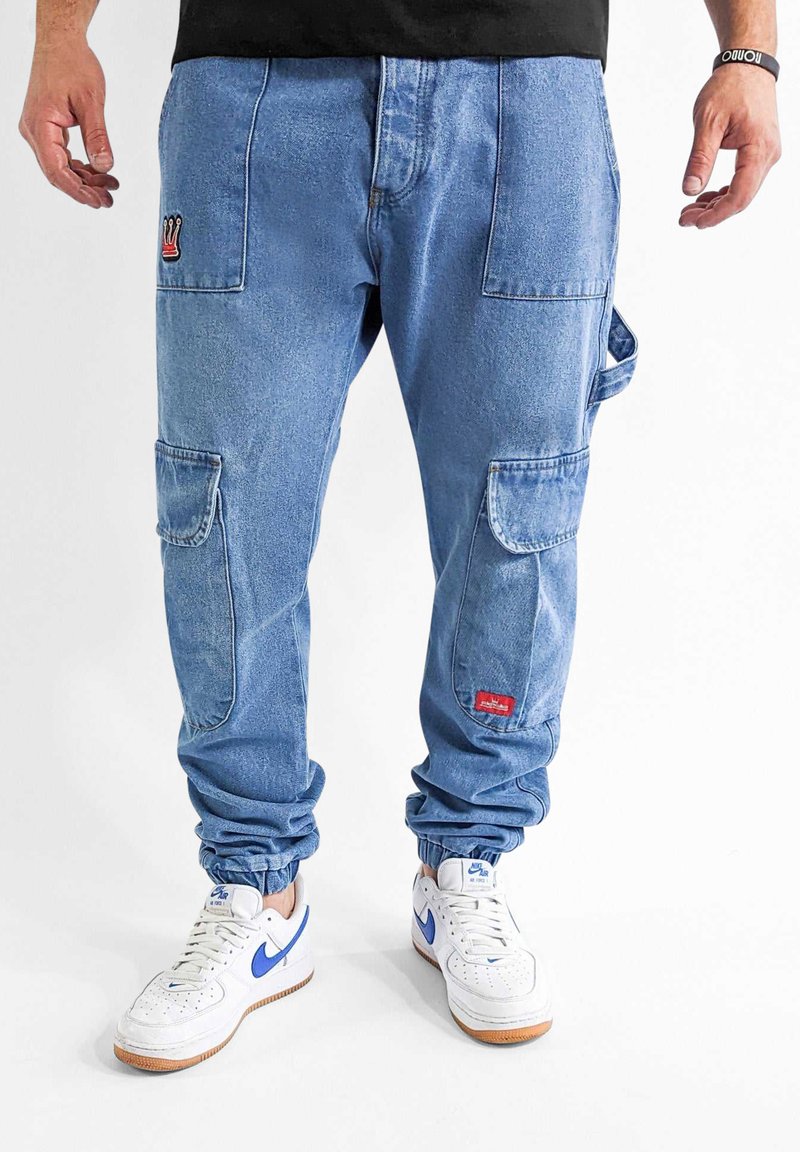 Helle blaue Cargohosen mit mehreren Taschen, einer gestickten Krone und Patch-Details, elastischen Bündchen und einem lässigen Schnitt. Getragen mit weißen Sneakers.