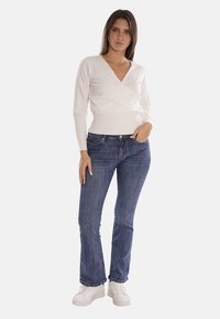 Maglione avvolgente bianco abbinato a jeans blu, con un design aderente e orlo a costine. Il modello indossa sneakers bianche su uno sfondo neutro.