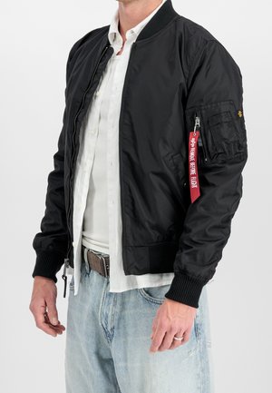Mand iført sort bomberjakke med rødt mærke "Remove Before Flight" på ærmet, hvid skjorte, lyseblå jeans og brun bælte.