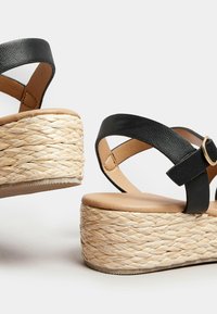 Mustat nahka wedge-sandaalit, joissa on kudottu raffia-alusta. Ominaisuuksiin kuuluvat ristiin menevät hihnat ja vaalea nahkainen pohja. Silkkinen pinta ja pyöreä muoto.