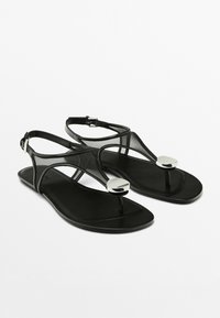 Černé sandály vyrobené z hladkého materiálu, s designem T-strap, lesklým stříbrným detailem a nastavitelným páskem kolem kotníku.