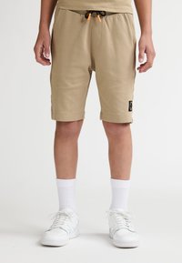 Beige bomuldsblandede shorts med snor i taljen, sidelommer og et sort logopatch på venstre ben. Bærer hvide sneakers og sokker.