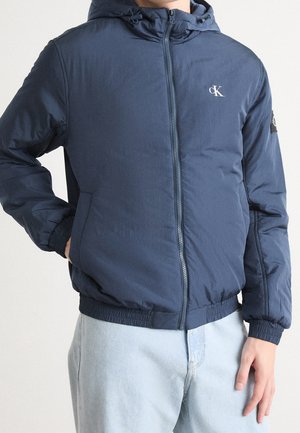 Person trägt eine marineblaue Kapuzenjacke mit Reißverschluss, elastischen Bündchen und Saum, die ein kleines weißes "cK"-Logo auf der Brust zeigt, sowie hellblaue Jeans.
