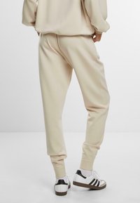 Beige joggebukser med tapered ben, elastiske mansjetter og en myk tekstur, kombinert med hvite sneakers med svarte striper og en teksturert hel.