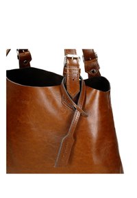 Sac à main en cuir marron avec des bretelles réglables et une étiquette en cuir décorative, mettant en valeur une texture lisse et des détails de boucle argentée.