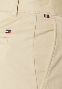 Beiga bomullbyxor har en slät yta med en sidficka och en liten broderad logotyp i röd, vit och blå nära fickan.