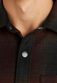 Camicia di flanella a quadri con tonalità rosso scuro e nero, bottoni automatici e tasca frontale per utilità. Tessuto morbido con vestibilità casual.