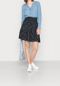 Chemise en denim bleu clair avec un col en V et des manches longues. Jupe noire à motif floral avec un ourlet volanté. Baskets blanches et sac à main bleu clair.