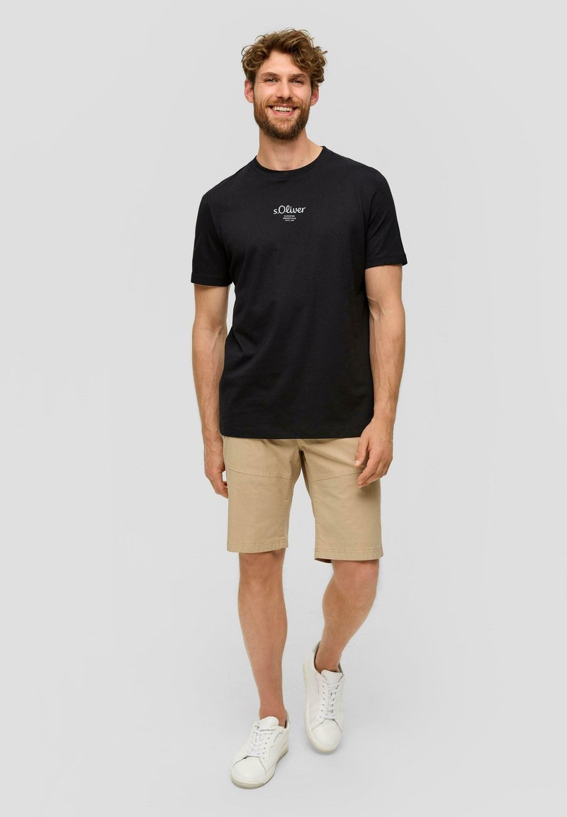 Maglietta nera con logo sul petto, abbinata a pantaloni corti beige e sneakers bianche. Tessuto di cotone, maniche corte, vestibilità casual.