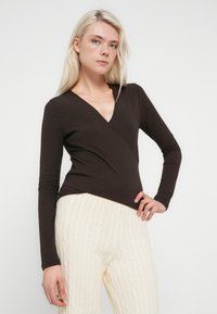 Bluză maro închis cu mâneci lungi, design cu decolteu în V suprapus, țesătură texturată, asortată cu pantaloni tricotati toronos cu culoarea crem deschis.