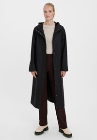 Black waterproof long coat with a hood, featuring snap-front closure, side pockets, and a relaxed fit, paired with brown pants and beige boots.