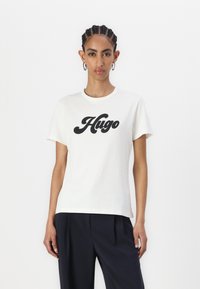 T-shirt en coton crème à manches courtes avec un grand logo noir "Hugo" sur la poitrine. Coupe simple et décontractée.