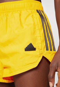 Κίτρινες σορτς από ελαφρύ ύφασμα με μαύρο λογότυπο Adidas και τρεις κατακόρυφες γκρίζες ρίγες στο πλάι. Οι πλαϊνές σχισμές βελτιώνουν την κινητικότητα.