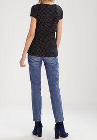 Kobieta w czarnej bluzce z krótkim rękawem w kropki, niebieskich dżinsach i granatowych botkach na obcasie, stojąca tyłem na jednolitym tle.