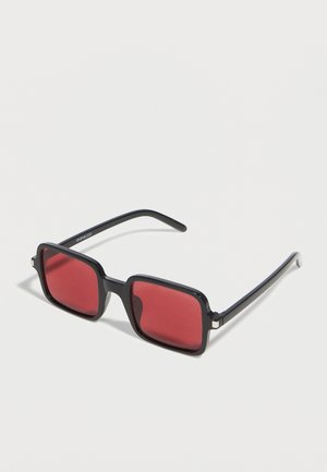 Lunettes de soleil à monture rectangulaire noire avec verres teintés rouges reposant sur une surface blanche.