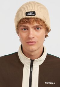 Beige ribaga ja musta ristkülikukujulise logo sildiga beanie, mida kannab inimene pruunis ja kreemis tõmblukuga jopega.