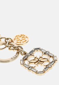 Brelok w złotym odcieniu z dekoracyjnym wisiorkiem o geometrycznym wzorze, ozdobiony prostokątnymi przezroczystymi kryształami. Dołączony jest okrągły charms.