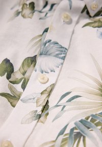 Camisa blanca con patrones de hojas tropicales en verde y azul, con botones en la parte delantera, fabricada con un tejido ligero y con un acabado texturizado.