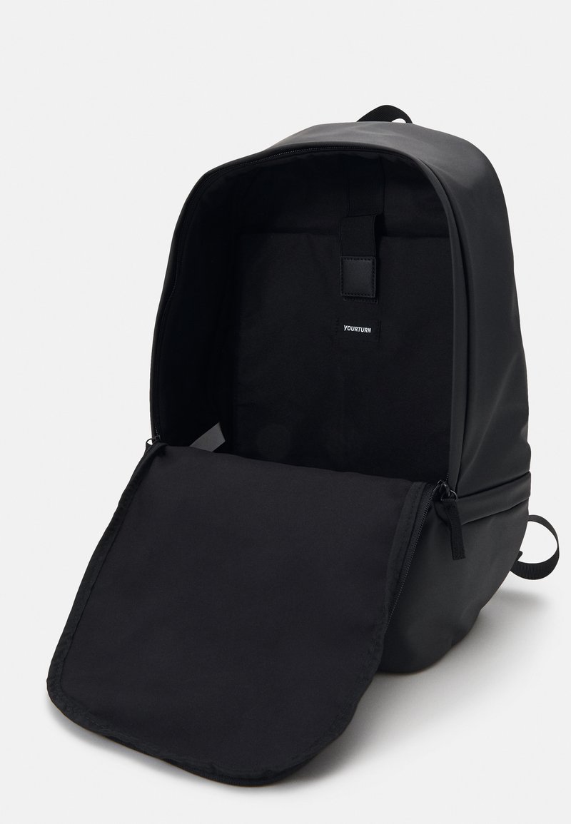 Mochila negra con un diseño elegante y minimalista; textura suave; solapa desabrochada que revela un interior espacioso con una pequeña etiqueta de marca.