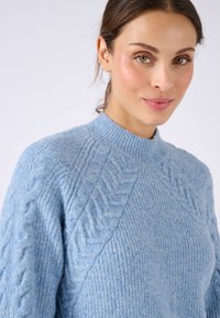 Maglione lavorato a maglia azzurro chiaro con colletto alto, caratterizzato da motivi a treccia in rilievo sulle spalle e sulle maniche, realizzato in morbido materiale peloso.