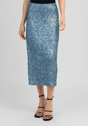 Femme portant une jupe midi bleue brillante à sequins, associée à des sandales noires à talons et à brides, ainsi qu'un top noir.
