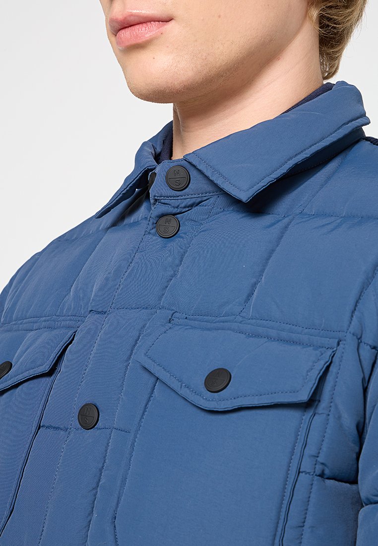 Giacca imbottita di colore blu con colletto con bottoni, dotata di due tasche al petto e una texture trapuntata. Sono visibili bottoni a pressione neri.