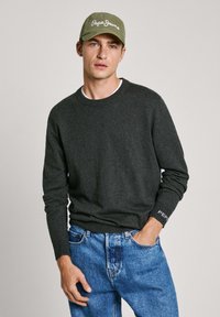 Šedá pletená mikina s dlouhými rukávy a kulatým výstřihem, doplněná světle modrými džíny. Zelená čepice s bílým textem dotváří outfit.