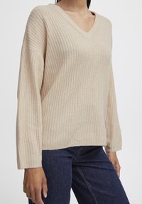 Beige bordás pulóver V-nyakkal, hosszú ujjal és laza szabással. Puha anyagból készült, textúrált, kellemesen meleg hatással.