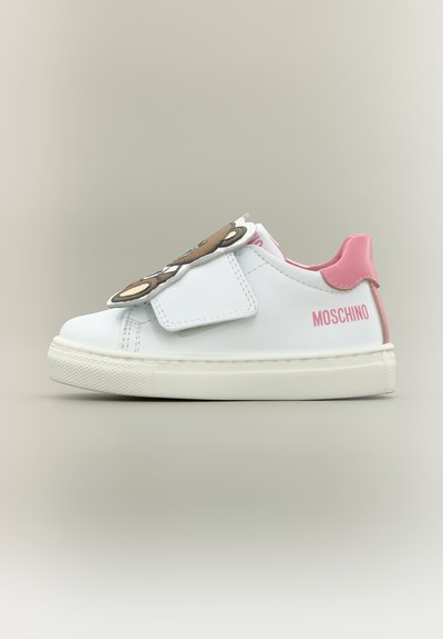 Bílá teniska s růžovou patní záložkou a logem "Moschino", se zdobením ve tvaru obličeje medvídka na pásku, boční pohled na neutrálním pozadí.