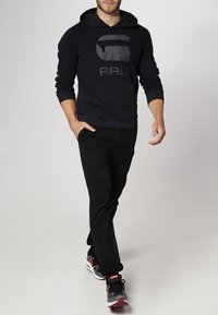 Felpa nera con un grande logo grigio "RAW", indossata con pantaloni neri e scarpe sportive con dettagli rossi. Tessuto morbido e vestibilità casual.