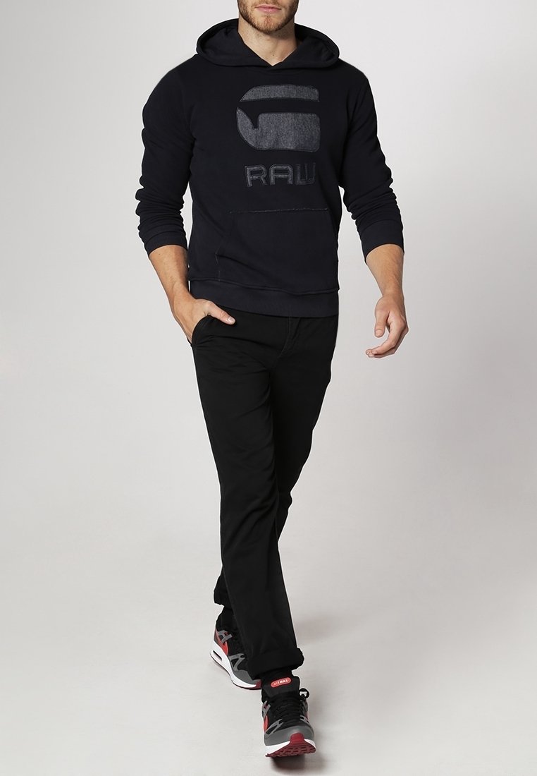 Felpa nera con un grande logo grigio "RAW", indossata con pantaloni neri e scarpe sportive con dettagli rossi. Tessuto morbido e vestibilità casual.