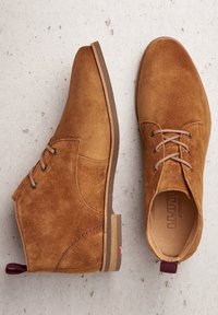 Botas de ante marrón hasta el tobillo con punta redonda, cordones y suela color beige. Presentan un tirador en el talón de color burdeos y sutiles acentos de costura.