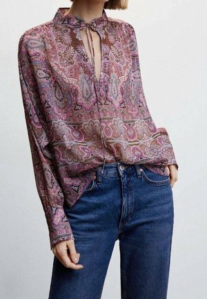 Kobieta w bluzce z długim rękawem i wzorem paisley z dekoltem typu keyhole oraz niebieskich dżinsach z wysokim stanem, stojąca z jedną ręką w kieszeni.