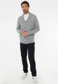 Mann trägt eine strukturierte graue Knopfpulloverjacke über einem weißen Hemd, schwarze Cargo-Hosen und weiße Sneakers und steht vor einem weißen Hintergrund.