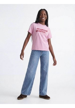 Smilende pige med flettet hår iført en lyserød Montreal Quebec t-shirt, vidde blå jeans og brune sneakers, stående på hvid baggrund.