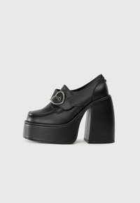 Melni platformas apavi ar noapaļotu purngali, ādas materiāls, vidusdaļas siksna ar sirds formas sprādzi un masīlu kvadrātveida papēdi.