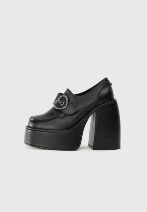 Sapatos pretos de plataforma com ponta arredondada, material em couro, fita na parte média do pé com uma fivela em forma de coração e salto quadrado robusto.
