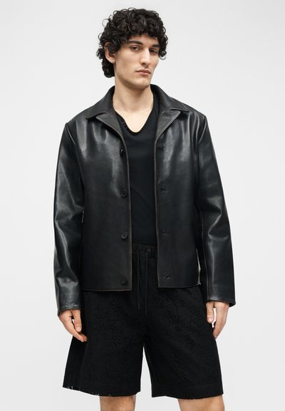 Νέος άνδρας με σγουρά μαλλιά, φορώντας μαύρο δερμάτινο σακάκι με κουμπιά πάνω από μαύρο πουκάμισο και μαύρο ανάγλυφο σορτς, στέκεται μπροστά σε ένα απλό φόντο.