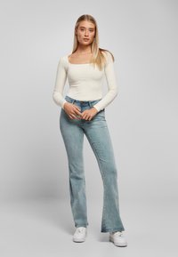 Top blanco de manga larga con escote cuadrado, combinado con jeans acampanados de color azul claro y zapatillas blancas. La modelo está de pie frente a un fondo gris liso.
