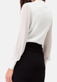 Femme portant un pull blanc avec des manches longues plissées et transparents, et une jupe noire taille haute, vue de dos.