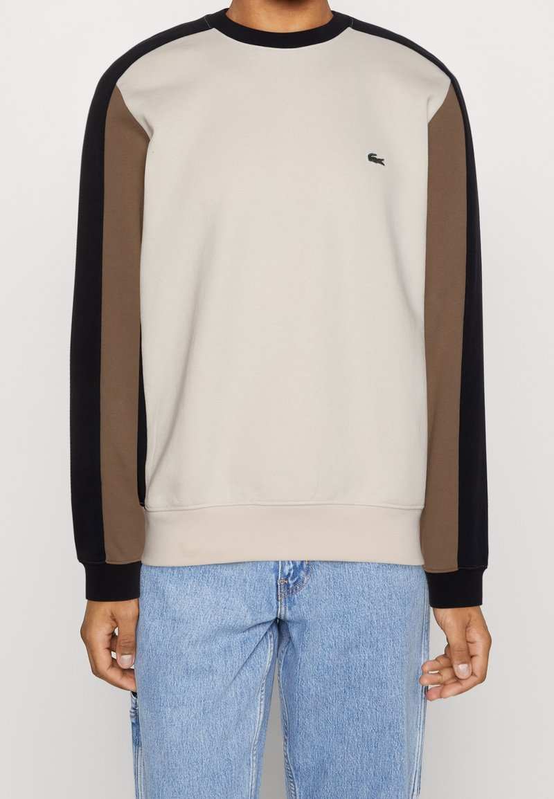 Hombre con sudadera beige de mangas largas negras y marrones, pequeño logo de cocodrilo en el pecho, y jeans azul claro.