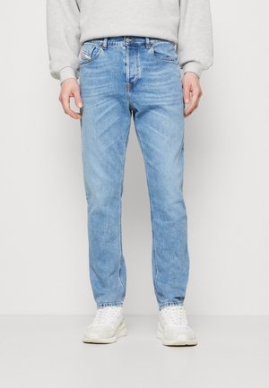 Mężczyzna ubrany w jasnoniebieskie jeansy o prostym kroju, białe trampki i jasnoszary sweter, stojący na jednolitym tle.