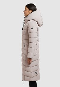 Svetlo roza dolg puffer plašč z visokim ovratnikom in kapuco, z prešitim vzorcem, stranskim zadrgo in črnim logotipom na robu.